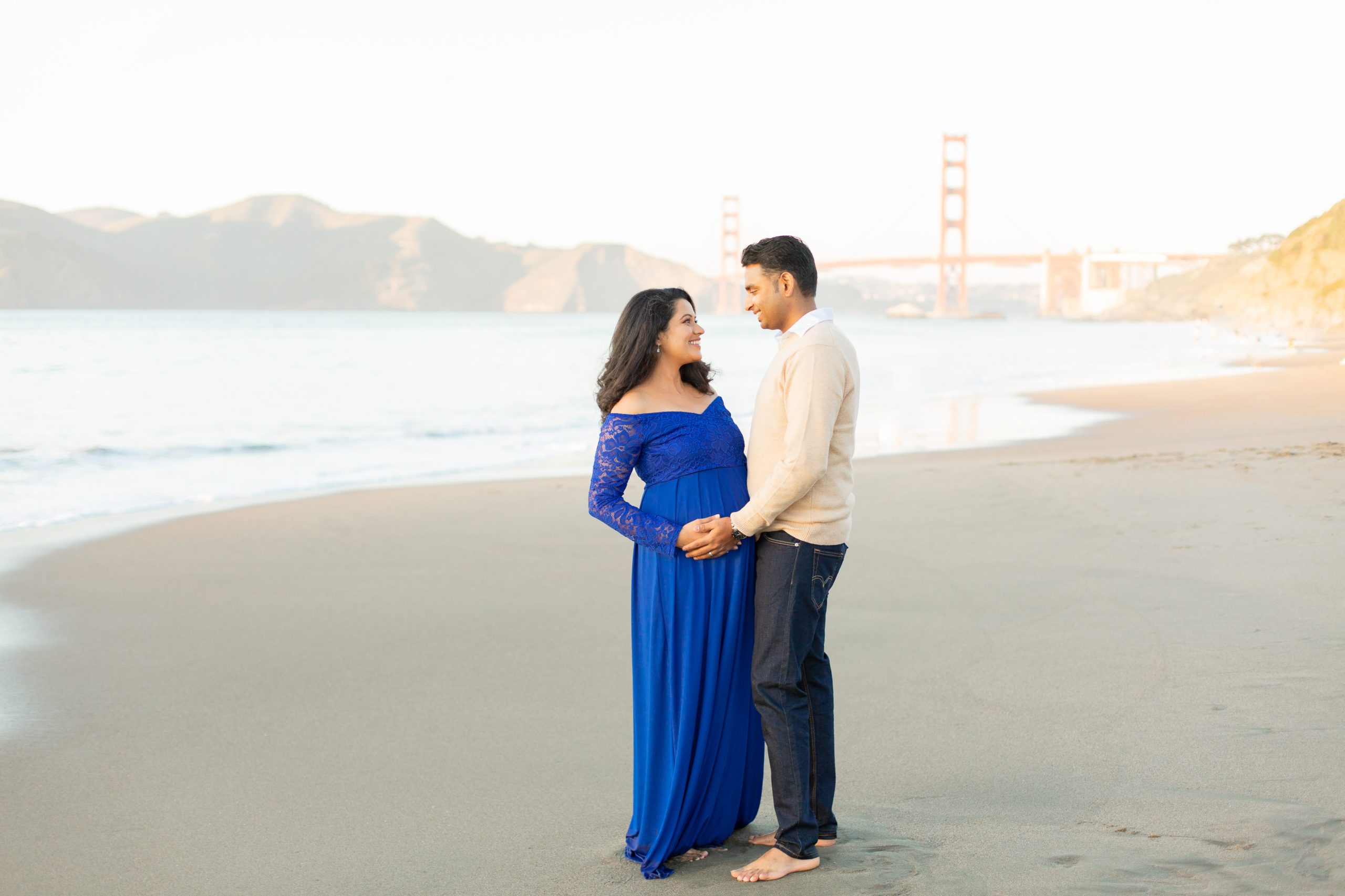 Pregnant woman and her husband embrace each other in on a beach in San Francisco with the Golden Gate Bridge in the background. The woman and man are Indian, both with dark hair. She is wearing a long blue dress and he is wearing a white shirt tan sweater and slacks.