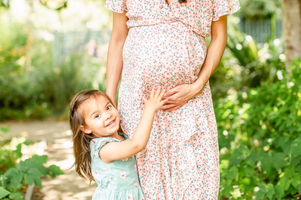 Woman expecting child poses while holding her belly, and her small daughter gives her pregnant stomach a squeeze