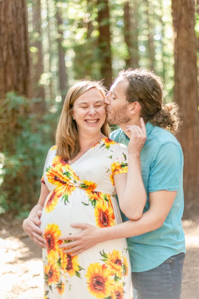 Couple expecting a child pose in the redwoods of the Bay Area, man kisses mother's cheek