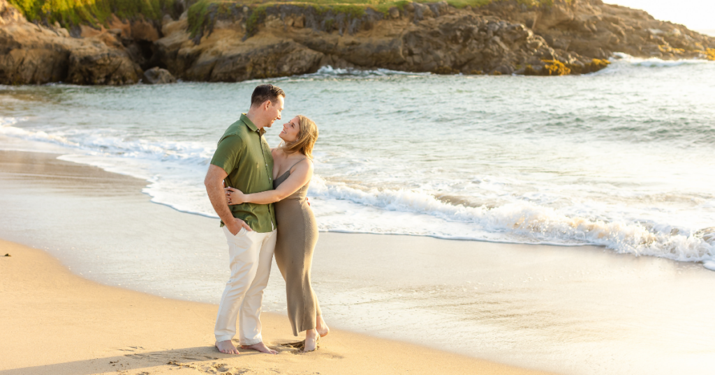 The couple shares a warm embrace with the beautiful beach in the background.